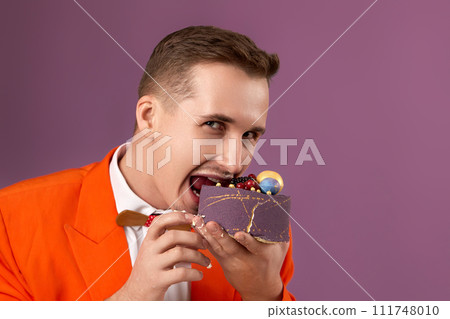 handsome birthday man in orange jacket bites cake on purple background 111748010