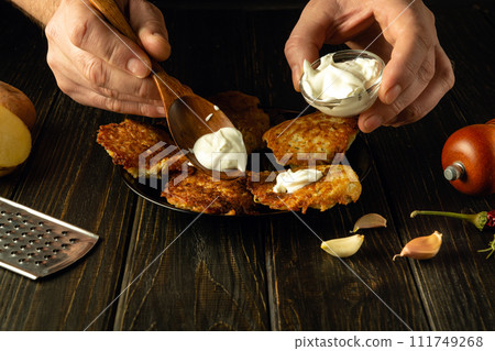 Male hands add sour cream with a spoon to fried potato pancakes on a plate. Concept of preparing delicious food at home. Advertising space 111749268