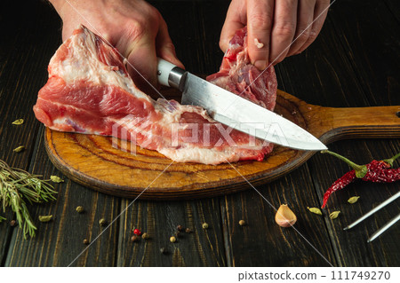 Close-up of a chef hands cutting raw veal meat with a knife before preparing dinner on the kitchen table in a restaurant 111749270