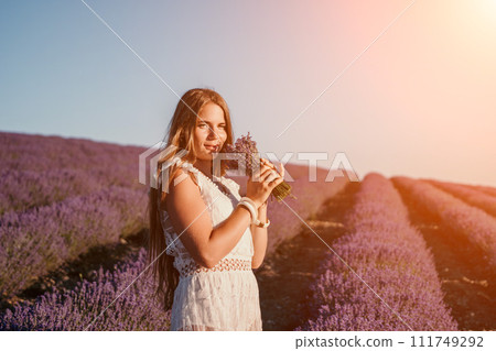 Woman lavender field. Happy carefree woman in a white dress walking in a lavender field and smelling a lavender bouquet on sunset. Ideal for warm and inspirational concepts in wanderlust and travel. Woman lavender field. Happy carefree woman in a white dress walking in a lavender field and smelling a lavender bouquet on sunset. Ideal for warm and inspirational concepts in wanderlust and travel. 111749292