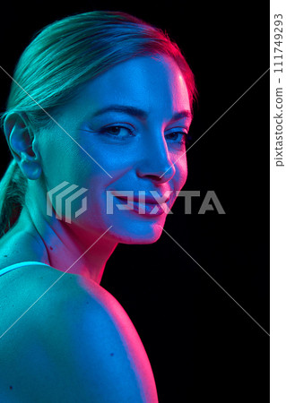 Young, pretty woman looking at camera over shoulder against black studio background in colorful neon light. Gel portrait. Young, pretty woman looking at camera over shoulder against black studio background in colorful neon light. Gel portrait. 111749293