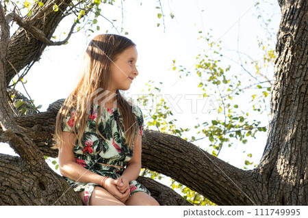 Cute five-year-old girl sitting on a tree in the summer in nature Cute five-year-old girl sitting on a tree in the summer in nature 111749995