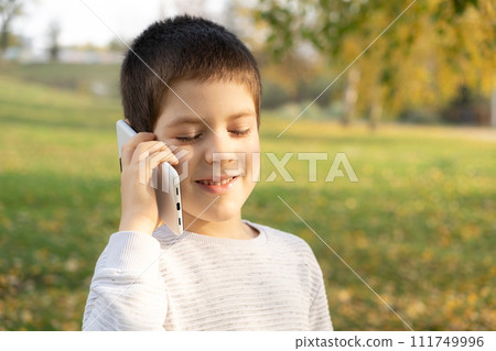 Handsome 6 year old boy talking on the phone while standing in park in autumn. Handsome 6 year old boy talking on the phone while standing in park in autumn. 111749996