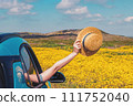 Feminine hand holding straw hat from the opened window of the car, among the yellow wild flowers. 111752040