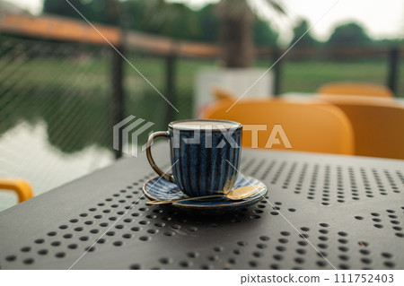 Coffee cappuccino with latte art in blue cup on iron table outdoors in city cafe. Beautiful foam, greenery ceramic cup.  111752403