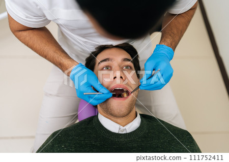 Portrait of handsome male patient lying in stomatology seat with open mouth during unrecognizable professional dentist examining teeth, with dental tool in dentistry clinic. Concept of stomatology. Portrait of handsome male patient lying in stomatology seat with open mouth during unrecognizable professional dentist examining teeth, with dental tool in dentistry clinic. Concept of stomatology. 111752411