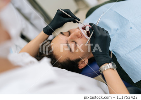 High-angle view of young man patient sitting in stomatology seat with open mouth during unrecognizable female dentist examining teeth, with dental tool in dentistry clinic. Concept of stomatology. 111752422