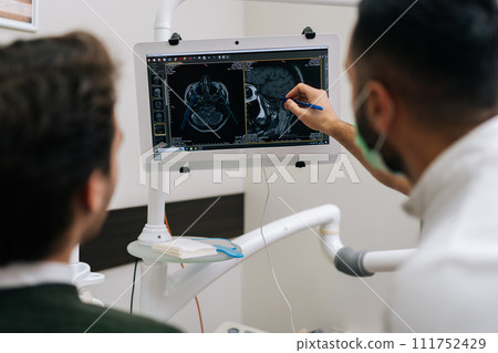 Rear view of male dentist pointing at teeth radiography on monitor to explain dentition diagnosis to young man patient with toothache. Dental expert showing x-ray scan results to man with cavity pain. 111752429
