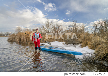 senior paddler wearing life jacket and drysuit is standing in water next to his stand up paddleboard on lake in Colorado in winter or early spring scenery senior paddler wearing life jacket and drysuit is standing in water next to his stand up paddleboard on lake in Colorado in winter or early spring scenery 111752534