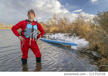 senior paddler wearing life jacket and drysuit is standing in water next to his stand up paddleboard on lake in Colorado in winter or early spring scenery 111752536