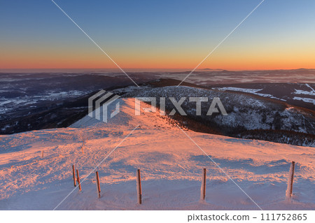 Winter, sunrise time, view from Snezka to pink mountain, krkonose mountain, Czech republic.. Along the road are wooden long bars, tourist markings for winter season Winter, sunrise time, view from Snezka to pink mountain, krkonose mountain, Czech republic.. Along the road are wooden long bars, tourist markings for winter season 111752865