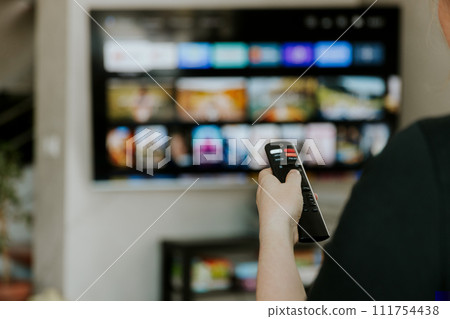 Hand holding remote control pointing to TV. Person watching smart television indoors. Blurred background. Colorful screen, copy space. Woman finger in foreground pressing button. Movie stream service Hand holding remote control pointing to TV. Person watching smart television indoors. Blurred background. Colorful screen, copy space. Woman finger in foreground pressing button. Movie stream service 111754438