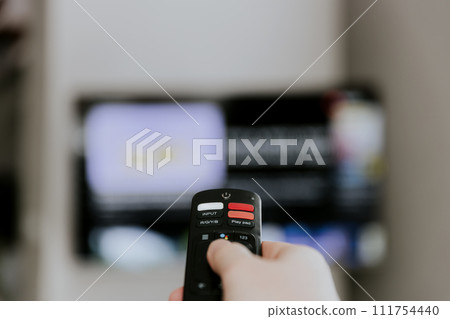 Hand holding remote control pointing to TV. Person watching smart television indoors. Blurred background. Colorful screen, copy space. Person browsing, looking for films online at home living room  111754440