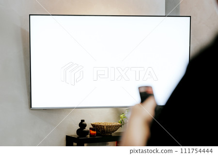 Hand holding remote control pointing to TV. Person watching smart television. Blurred background. White empty mockup screen, copy space. Manage appliances, elements of a smart home control panel Hand holding remote control pointing to TV. Person watching smart television. Blurred background. White empty mockup screen, copy space. Manage appliances, elements of a smart home control panel 111754444