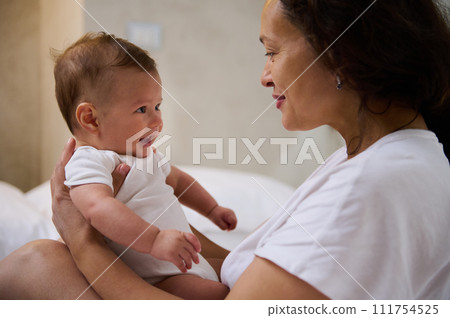 Loving mom sitting on the bed, holding her baby boy, communicating and smiling to him, establishing emotional contact 111754525
