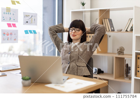 Caucasian woman in formal wear relaxing in office chair with arms behind head. 111755784