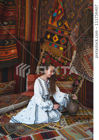 beautiful girl in a white dress with a teapot in a traditional turkish interior with many carpets on the wall beautiful girl in a white dress with a teapot in a traditional turkish interior with many carpets on the wall 111756407