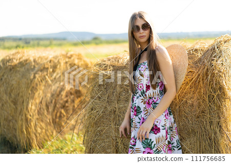 beautiful glamorous girl in a dress and a straw hat on a field near a haystack. 111756685