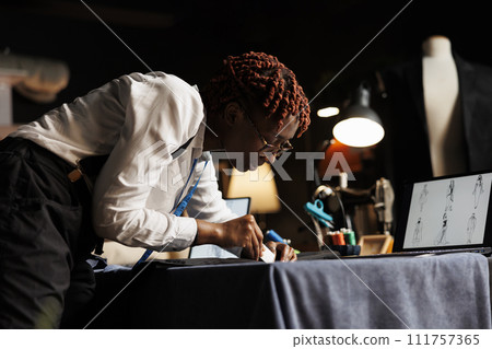 Hip level shot of african american tailor carefully checking suit blazer seam to make sure it fits customer. Experienced seamstress manufacturing premium quality elegant garment 111757365