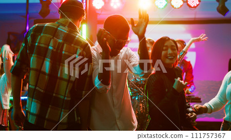 Diverse friends dancing on live music, feeling cheerful on dance floor at nightclub. Young people having fun with performance at discotheque, doing funky dance moves under stage lights. Tripod shot. 111757367