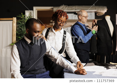 Suitmaker taking customer measurements for bespoken handmade business suit while elderly needleworker does adjustments on sartorial garment. Teamworking tailors manufacturing premium quality clothing 111757369
