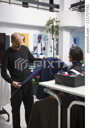 Arab man examining apparel size and style while talking with clothing store consultant. Shopping center fashion department employee giving buyer advice in selecting formal jacket 111757381