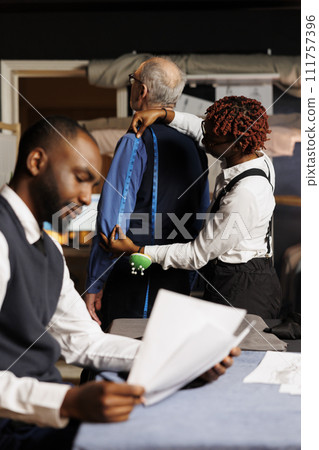 Teamworking tailors helping senior client find perfectly fitting bespoke business suit. Cheerful couturiers taking elderly gentleman measurements during sartorial customization comission process Teamworking tailors helping senior client find perfectly fitting bespoke business suit. Cheerful couturiers taking elderly gentleman measurements during sartorial customization comission process 111757396