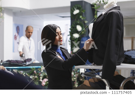 Clothing store asian woman assistant examining jacket on mannequin while putting on suit on mannequin. Boutique seller creating stylish formal outfit to showcase apparel brand 111757405