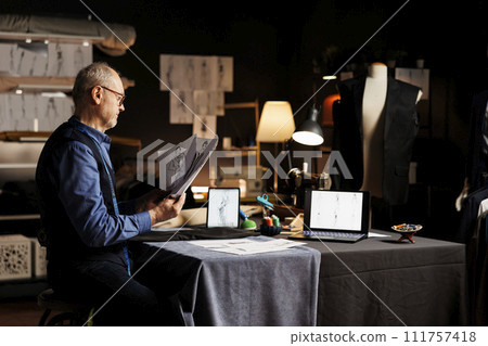 Couturer in atelier shop workspace comparing handdrawn sketch designs for sartorial collection. Elderly tailor trying to pick perfect bespoken attire for upcoming fashion runway show Couturer in atelier shop workspace comparing handdrawn sketch designs for sartorial collection. Elderly tailor trying to pick perfect bespoken attire for upcoming fashion runway show 111757418