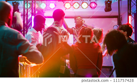 Multiethnic group of people partying on dance floor, enjoying electronic funky music from DJ mixing station. Cheerful adults having fun at nightclub, dancing together at celebration. Handheld shot. Multiethnic group of people partying on dance floor, enjoying electronic funky music from DJ mixing station. Cheerful adults having fun at nightclub, dancing together at celebration. Handheld shot. 111757428