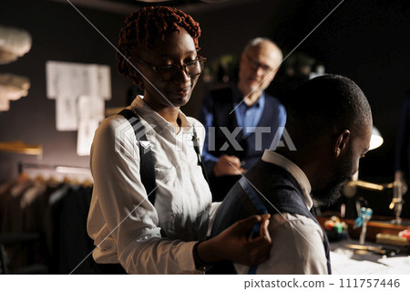 Aspiring seamstress apprentice learning craftmanship from experienced elderly suitmaker in tailoring studio workspace. Couturier doing bespoken sartorial fit sizing under master guidance 111757446