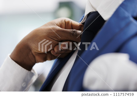 African american man arms fixing tie on male mannequin in fashion showroom. Boutique seller wearing formal suit on model and adjusting jacket, showcasing clothes to customer in mall 111757459
