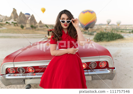 beautiful girl in retro style posing near a vintage red cabriolet car on background of balloons in Cappadocia. 111757546