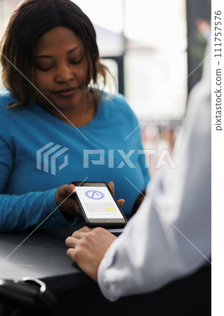 African american shopper buying clothes with phone nfc payment, using smartphone at pos terminal in modern boutique store. Woman paying for fashionable casual wear and accessories at counter. Close up 111757576