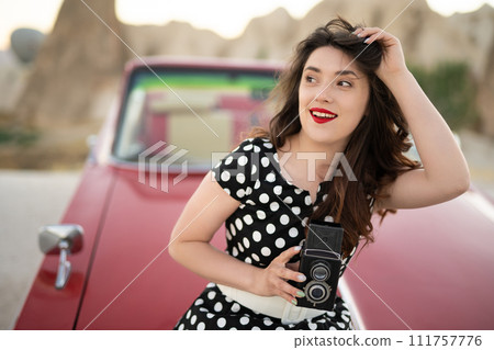 beautiful girl in retro style posing near vintage red cabriolet car with old film camera in hands. beautiful girl in retro style posing near vintage red cabriolet car with old film camera in hands. 111757776