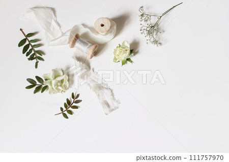 Floral wedding, birthday table composition. Gypsophila, rose flowers, green lentisk leaves and silk ribbons with wooden spools isolated on white table background. Flat lay, top view. Copy space, no Floral wedding, birthday table composition. Gypsophila, rose flowers, green lentisk leaves and silk ribbons with wooden spools isolated on white table background. Flat lay, top view. Copy space, no 111757970