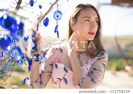 a portrait of a beautiful girl in Turkish hangs many amulets nazar in the shape of an eye near a tree 111758234
