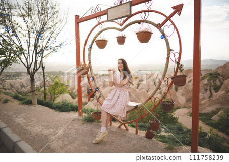 Girl sitting on a decorated heart-shaped bench on a viewpoint and admiring view of Cappadocia Girl sitting on a decorated heart-shaped bench on a viewpoint and admiring view of Cappadocia 111758239