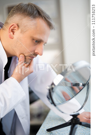 the man is a doctor, beautician looks at his skin in the mirror the man is a doctor, beautician looks at his skin in the mirror 111758253