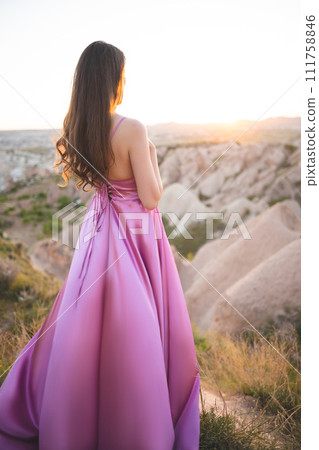 a beautiful girl in a long pink dress poses at sunset in the mountains of Cappadocia 111758846