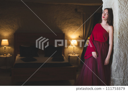 a girl in a red evening dress poses in the interior of a hotel bedroom 111758879