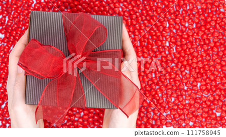 Female's hands holding Christmas gift box decorated with red ribbon bow on red candy background. Christmas and New Year banner. Christmas shopping, Boxing day, pepermint candies. Female's hands holding Christmas gift box decorated with red ribbon bow on red candy background. Christmas and New Year banner. Christmas shopping, Boxing day, pepermint candies. 111758945
