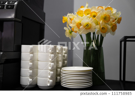 Many clean white cups and plates on the table near the coffee maker and vase with daffodils 111758985