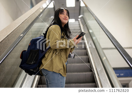 A positive young Asian female solo traveler is on an escalator in the airport. 111762820