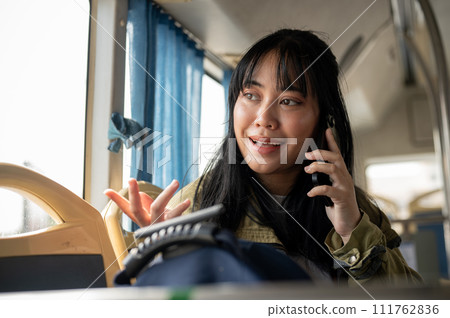 A happy Asian woman is on the bus, looking out the window while talking on the phone. A happy Asian woman is on the bus, looking out the window while talking on the phone. 111762836