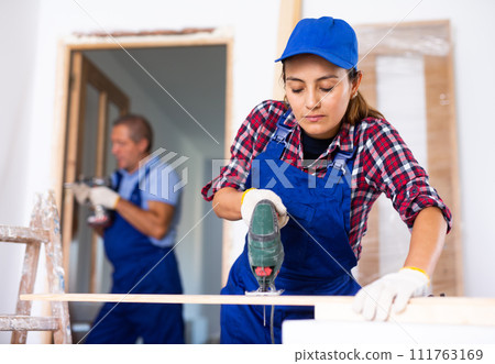 Focused Latin female in uniform using a jigsaw machine on wooden plank at home 111763169