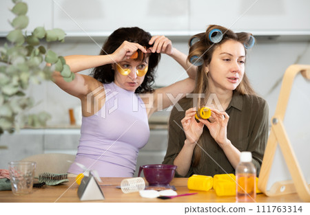 Female friends curling hair with rollers by tabletop mirror 111763314
