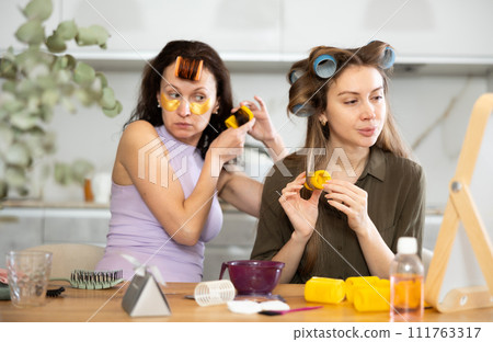 Female friends curling hair with rollers by tabletop mirror Female friends curling hair with rollers by tabletop mirror 111763317