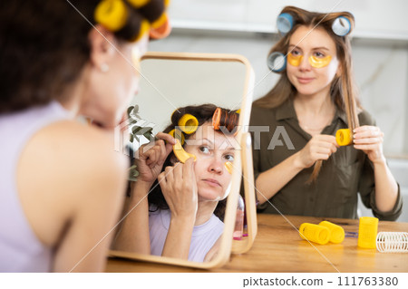 Woman in hair rollers applying eye patches in front of mirror with friend 111763380