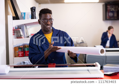 Excited middle-aged African American male typographer in uniform showing large format paper in a roll in the typography Excited middle-aged African American male typographer in uniform showing large format paper in a roll in the typography 111763403